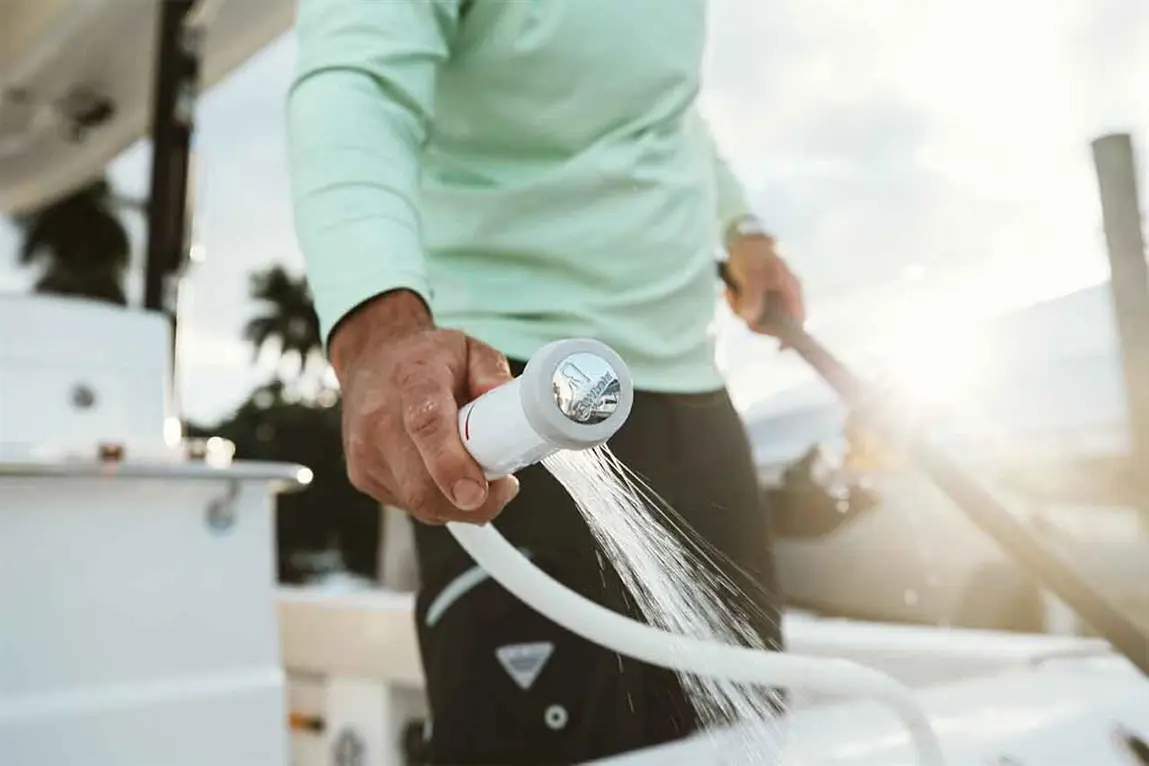 A man in a light green shirt washes the deck of a boat using a hose with water spraying from the nozzle, sunlight shining behind.